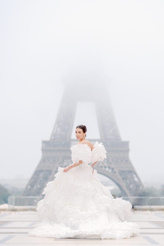 Paris pre-wedding session in front of the Eiffel Tower while it is covered in fog with bride in full train layered gown with separate off the shoulder full puffed sleeves and groom in a navy blue suit photographed by Italy destination photographer